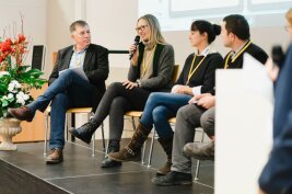 Mehrere Personen sitzen auf Stühlen auf einem Podium, eine Frau spricht in ein Mikrofon.