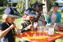 Vier Schulkinder an einem Aktionstisch löffeln aus Gläsern. Die Kinder tragen LfL-Mützen.