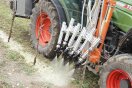 Ein Traktor mit seitlich angebrachten Düsenapparaten spritzt weißes und gelbes Material auf Boden unter Weinreben